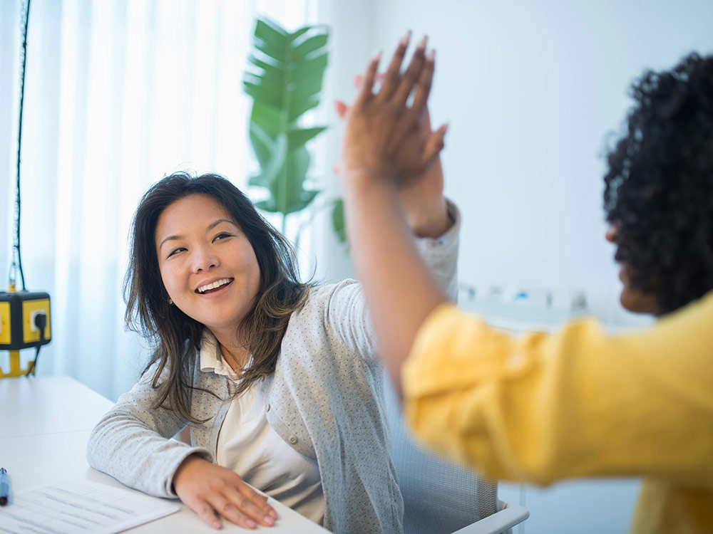 Two women high five at a desk, symbolizing emotional resilience and support in a collaborative environment